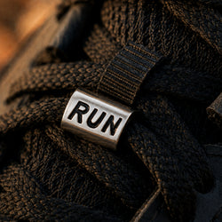 A close-up of a silver metal shoe charm engraved with the word 'RUN', attached to black athletic laces on a sneaker.