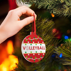 Person holding a round Christmas ornament with a red ribbon hanger, featuring the word 'VOLLEYBALL' in red letters over a volleyball graphic on a white band, surrounded by red, green, and white holiday patterns with a Christmas tree in the background.