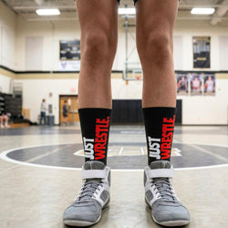 Person wearing 'Just Wrestle' socks and gray wrestling shoes on a gym floor.