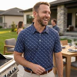 Man wearing a blue soccer patterned polo shirt standing outdoors with a house and people in the background