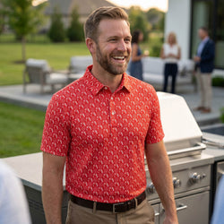 Man wearing a red wrestling patterned polo shirt standing outdoors near a grill with people in the background.