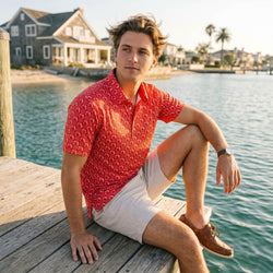 Teen in a red wrestling patterned polo shirt and beige shorts sitting on a dock by water