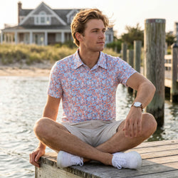 Teen sitting on a dock by a body of water wearing a wrestling patterned polo shirt and beige shorts.