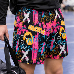 A close-up of a person wearing black athletic shorts with a vibrant multicolor graffiti-style wrestling graphic print, standing on a blue court.