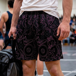 A person standing in a gym wearing black athletic shorts with a pink line-art wrestling graphic pattern, holding a black and gray duffel bag. Other athletes and a blurred indoor court are visible in the background.